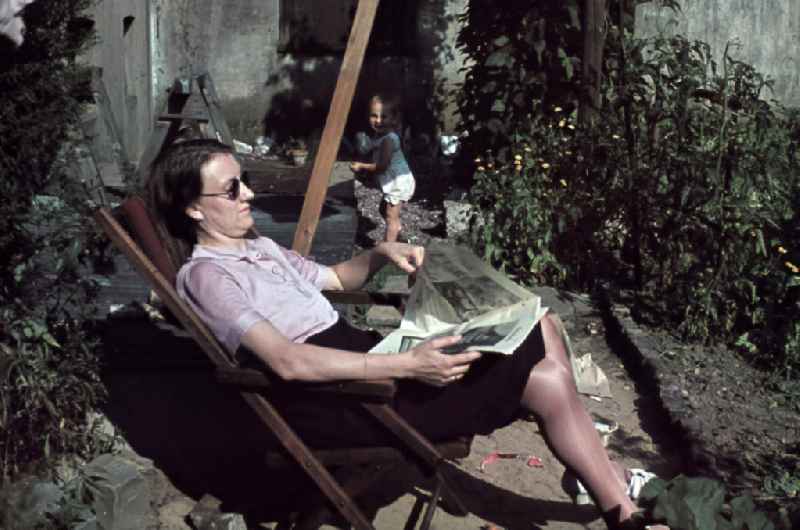 Kleinkind spielt im Garten, die Mutter sitzt im Liegestuhl und liest eine Zeitung. Plying infant in a garden. The mother is sitting in a deck chair and read a newspaper.
