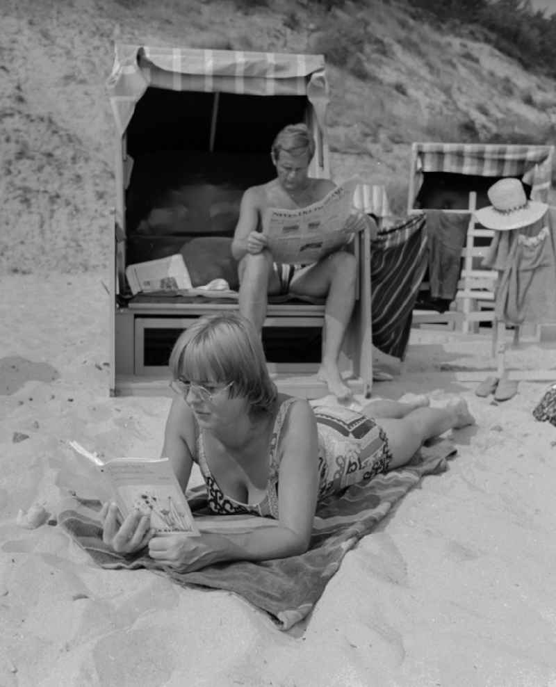 Tourists on the beach in Ueckeritz in Mecklenburg-Western Pomerania in the field of the former GDR, German Democratic Republic. A man sitting in a beach chair and reading a newspaper. A woman with glasses lying on a towel reading a book