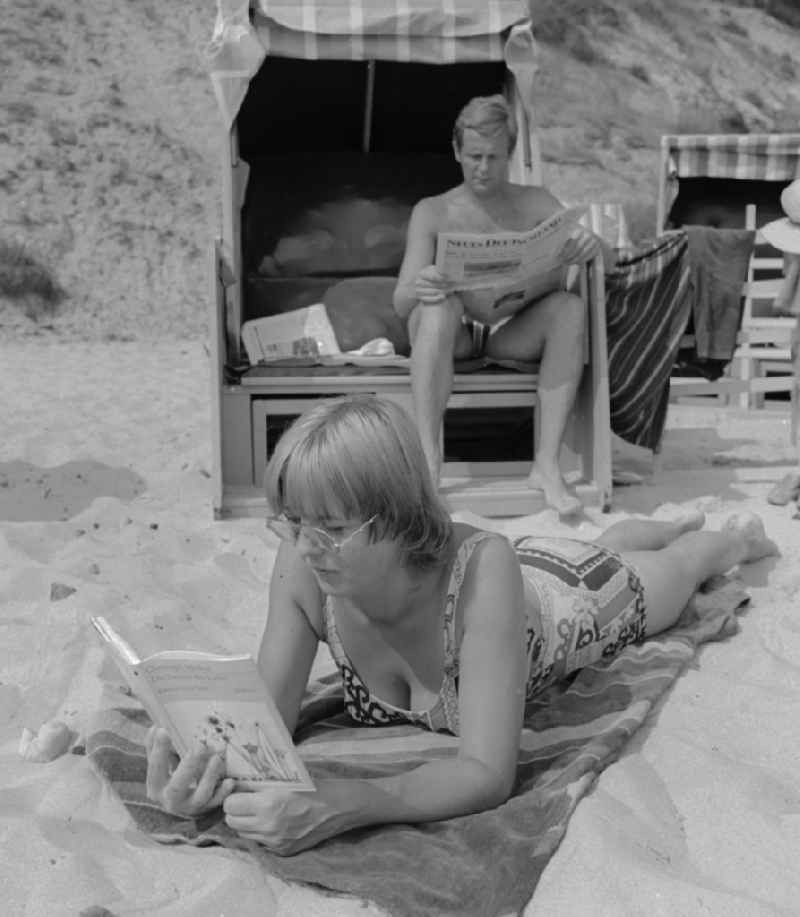 Tourists on the beach in Ueckeritz in Mecklenburg-Western Pomerania in the field of the former GDR, German Democratic Republic. A man sitting in a beach chair and reading a newspaper. A woman with glasses lying on a towel reading a book