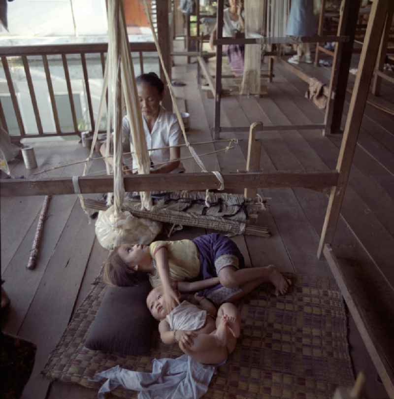 Eine Weberin bei ihrer Arbeit, während die Kinder daneben auf einer Matte liegen in einer Weberei in Vientiane, der Hauptstadt der Demokratischen Volksrepublik Laos.