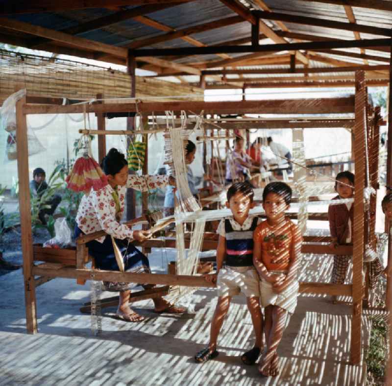 Frauen bei ihrer Arbeit, während die Kinder daneben spielen in einer Weberei in Vientiane, der Hauptstadt der Demokratischen Volksrepublik Laos.