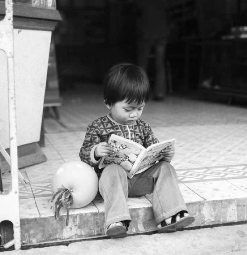 Ein kleiner Junge sitzt am Straßenrand auf einer Bordsteinkante und betrachtet ein Comicbuch in Vientiane, der Hauptstadt der Demokratischen Volksrepublik Laos.