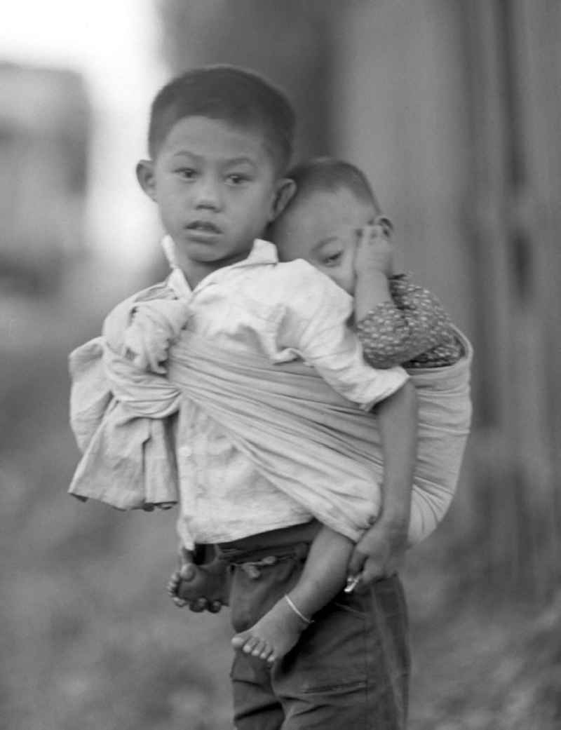 Ein Junge trägt sein kleines Geschwisterkind in einem Tragetuch auf seinem Rücken auf einer Straße in Vientiane, der Hauptstadt der Demokratischen Volksrepublik Laos.