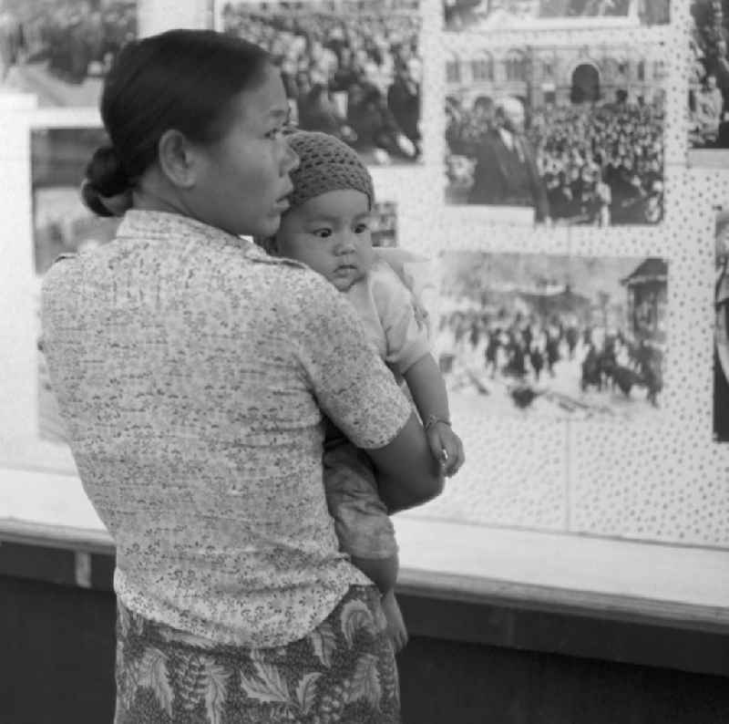 Ein Jahr nach der Gründung der Volksrepublik Laos im Dezember 1975 betrachtet eine Frau mit ihrem Baby auf dem Arm in einem Schaukasten ausgestellte Lenin-Bilder von der russischen Oktoberrevolution in Vientiane in der Demokratischen Volksrepublik Laos.