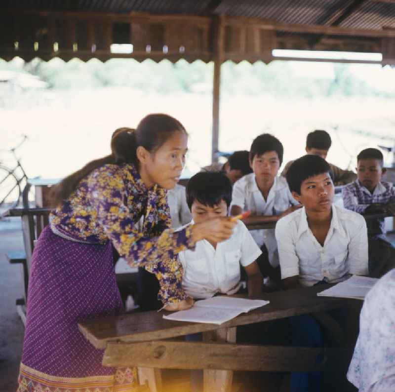 Eine Lehrerin unterrichtet ihre Schüler im Lesen und Schreiben in einer Schule in einem Dorf in der Demokratischen Volksrepublik Laos. Nach der Gründung der Volksrepublik Laos im Dezember 1975 wurde das Schulsystem von der neuen kommunistischen Regierung in Laos wieder aufgebaut, um dem in der Bevölkerung weit verbreiteten Analphabetismus entgegenzuwirken.