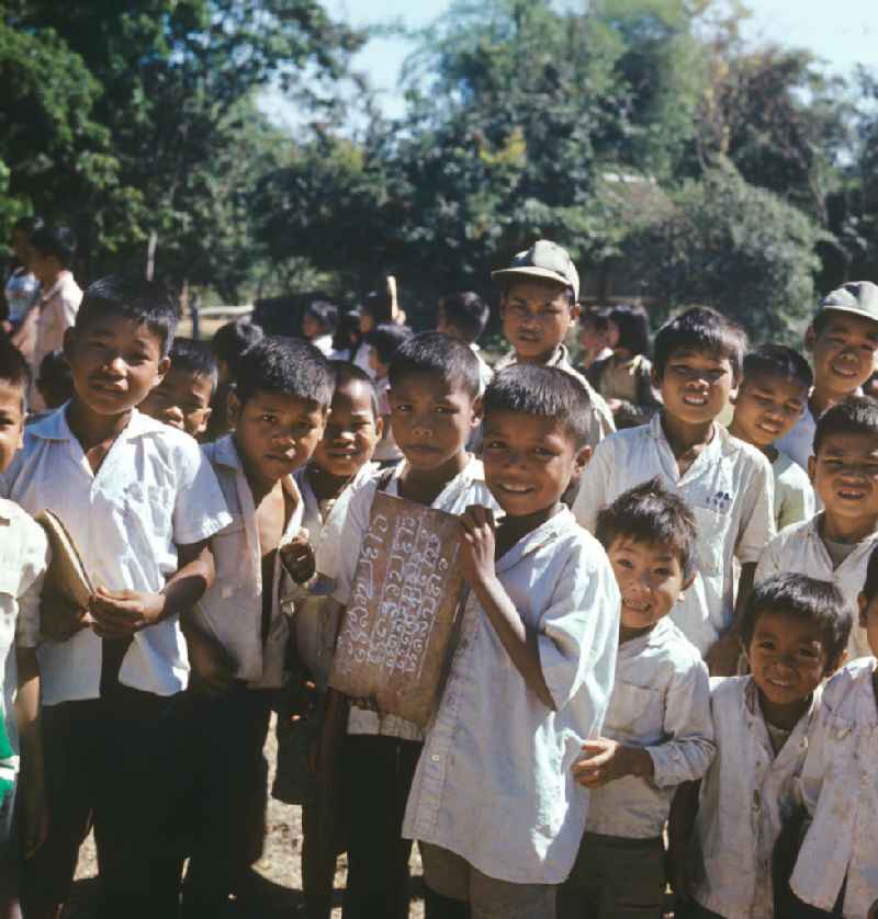 Ein Junge präsentiert dem Fotografen eine Schreibtafel auf dem Hof einer Schule in einem Dorf in der Demokratischen Volksrepublik Laos. Nach der Gründung der Volksrepublik Laos im Dezember 1975 wurde das Schulsystem von der neuen kommunistischen Regierung in Laos wieder aufgebaut, um dem in der Bevölkerung weit verbreiteten Analphabetismus entgegenzuwirken.