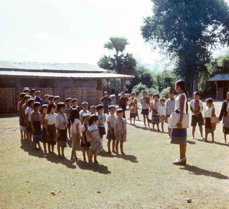Kinder versammeln sich auf dem Hof einer Schule in einem Dorf in der Demokratischen Volksrepublik Laos. Nach der Gründung der Volksrepublik Laos im Dezember 1975 wurde das Schulsystem von der neuen kommunistischen Regierung in Laos wieder aufgebaut, um dem in der Bevölkerung weit verbreiteten Analphabetismus entgegenzuwirken.
