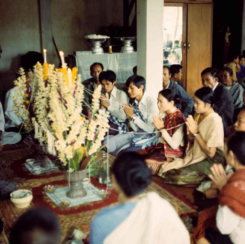 Feierliches Ritual in Anwesenheit buddhistischer Mönche bei einer Hochzeit in einem Dorf in der Demokratischen Volksrepublik Laos. Bis zur Ausrufung der Volksrepublik Laos am 2. Dezember 1975 bestimmte der Theravada-Buddhismus die kulturelle Entwicklung im Land. Nachdem die anfängliche Unterdrückung der traditionellen buddhistischen Bräuche durch die neuen kommunistischen Machthaber mißlang, fand Laos in den folgenden Jahrzehnten einen eigenen Weg der Koexistenz von Buddhismus und Sozialismus.