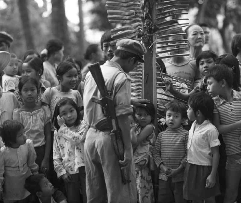 Ein Milizionär streichelt einem Mädchen über den Kopf, das zusammen mit anderen Kindern und Familien in Vientiane auf die Rückkehr der 'umerzogenen' Männer und Frauen von der Frauen- und Männerinsel in der Demokratischen Volksrepublik Laos wartet. Das Nam Ngum Reservoir entstand mit der Fertigstellung des gleichnamigen Staudamms im Jahr 1971. Die im See liegenden Inseln dienten in den 7