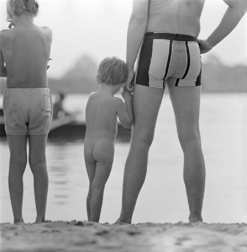 Father with son by the hand standing on the shore and water area of ??Lake Riewendsee with bathers in Wachow, Brandenburg in the area of ??the former GDR, German Democratic Republic