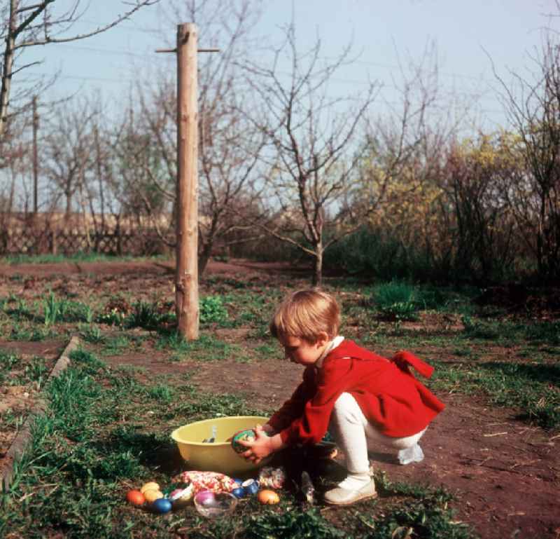 A small child in a red dress with the Easter egg look in the garden in Zschopau in the federal state Saxony in the area of the former GDR, German democratic republic
